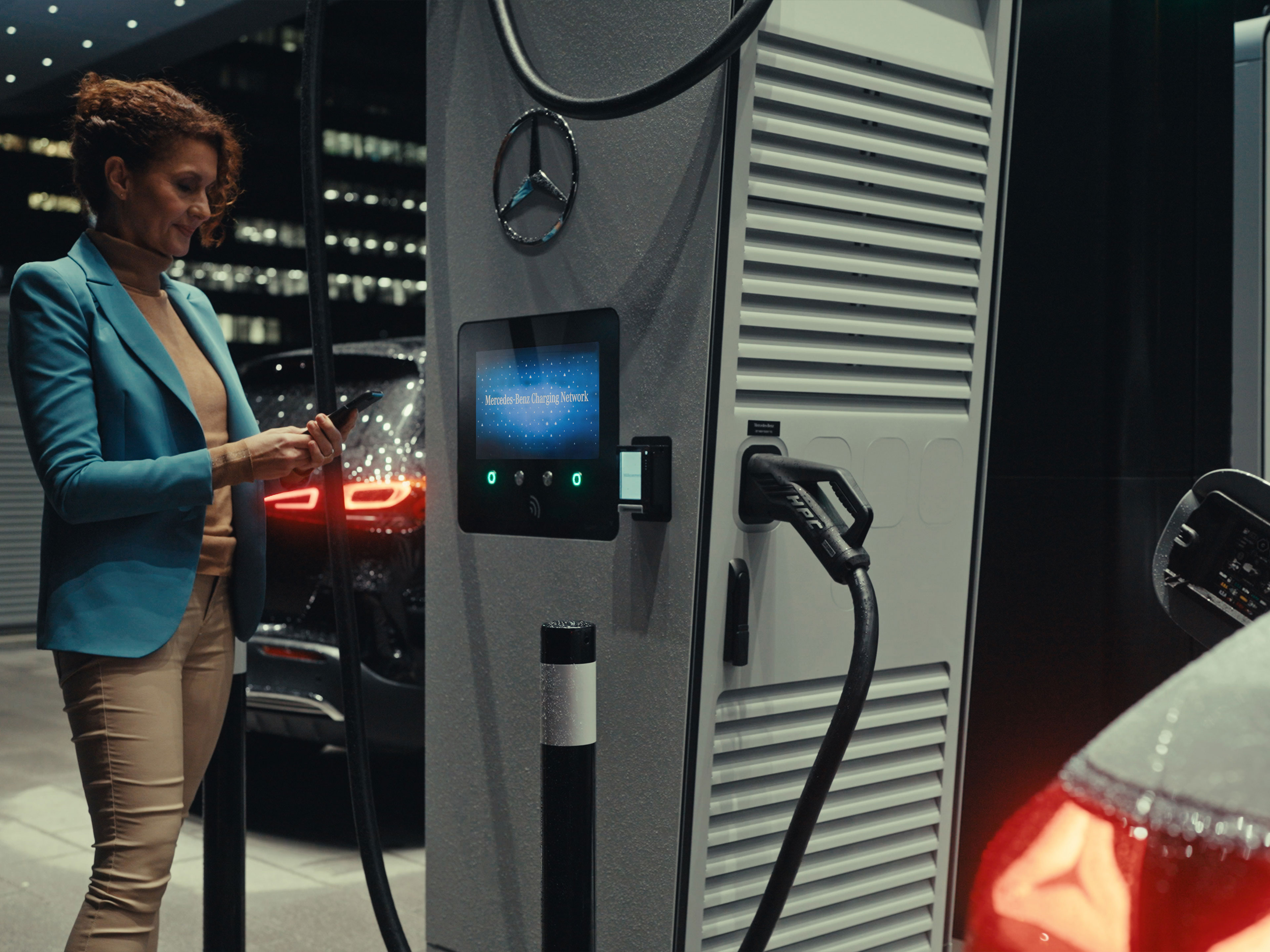 Mercedes-Benz App | Charging on the go | Mercedes-Benz A woman holding a smartphone in her hand, standing next to a charging station.