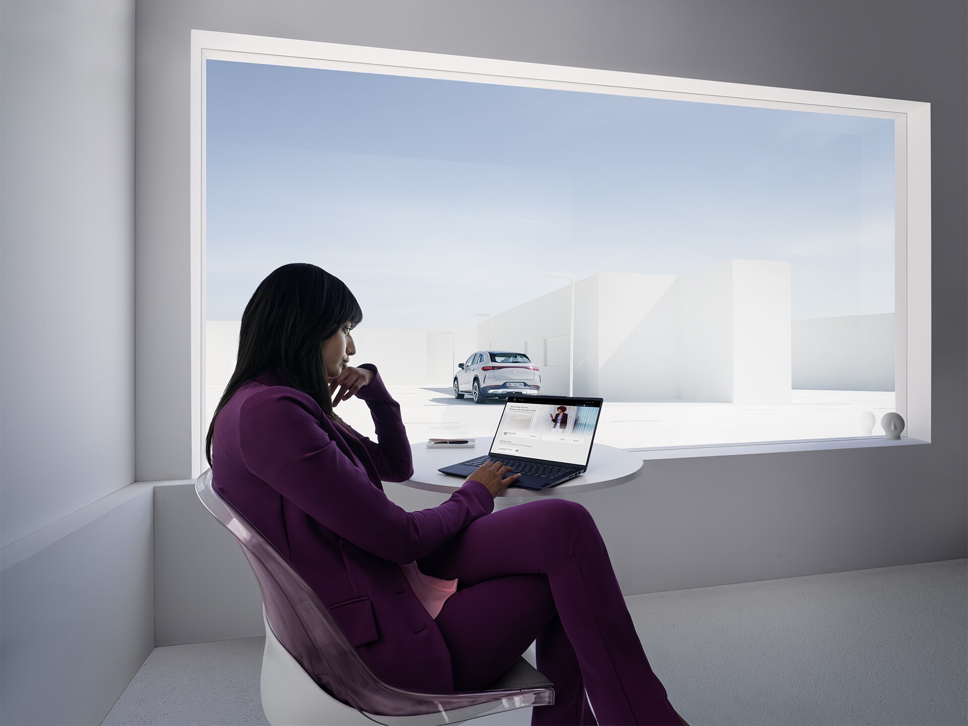 A woman working at a laptop sits in a white office in front of a large window.