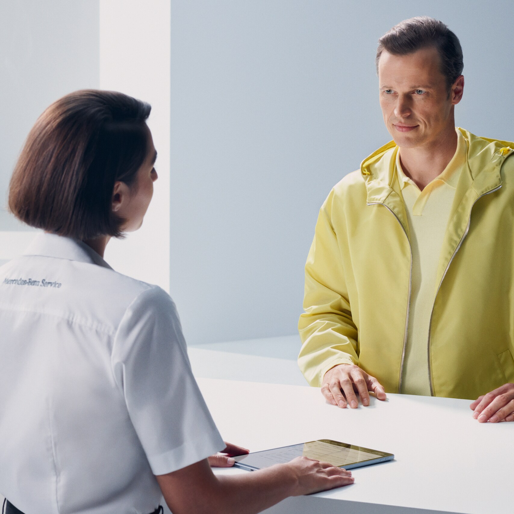 A customer leaning at the counter turns expectantly to face a Mercedes-Benz employee.