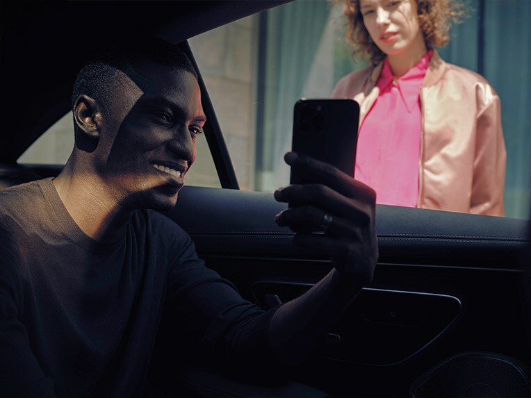 A man sitting in a Mercedes-Benz shows his mobile phone to a woman standing outside