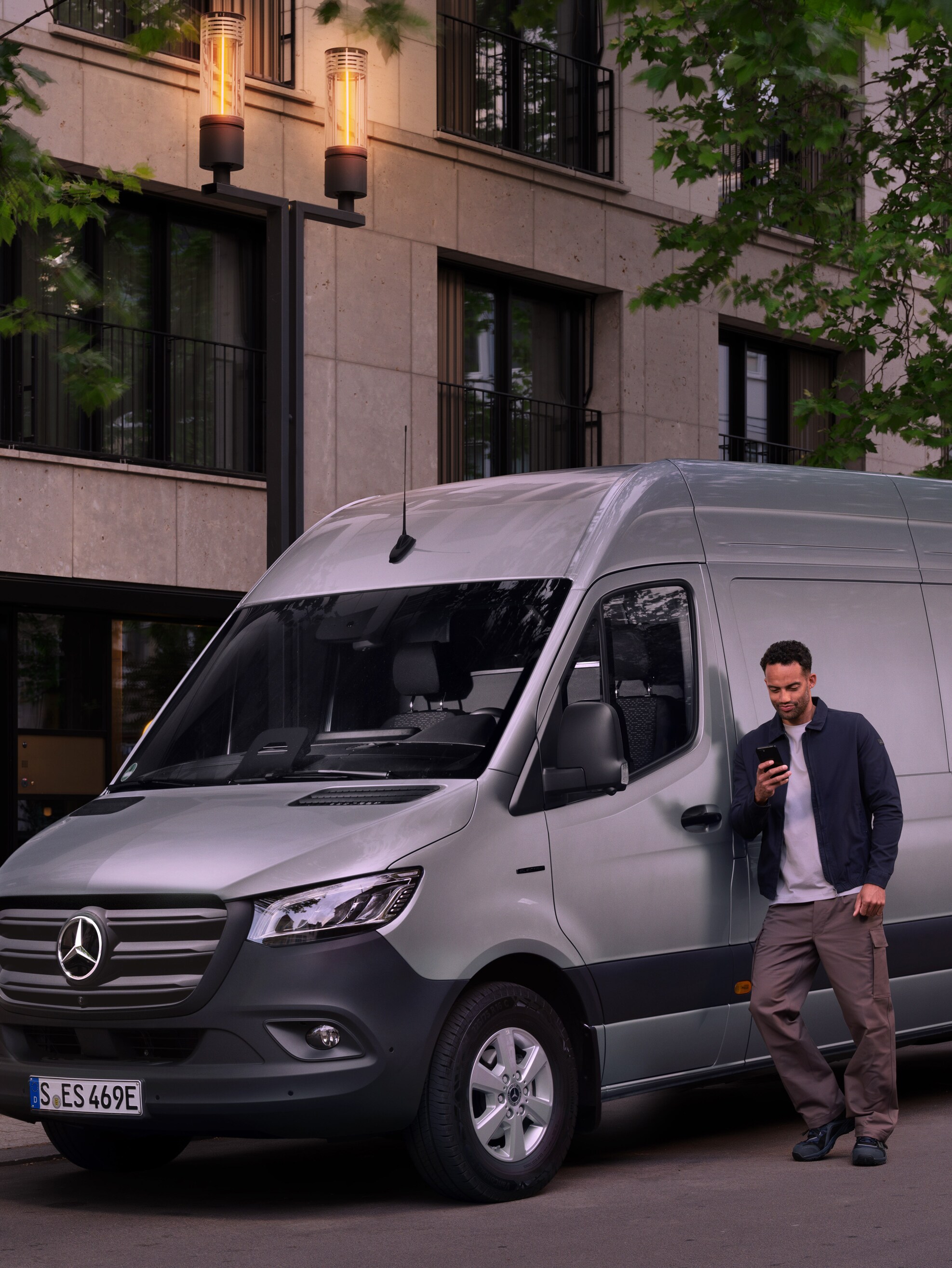 Man leaning on a silver Mercedes-Benz Sprinter van and using his smartphone – speedy assistance from Mercedes Benz Customer Care.