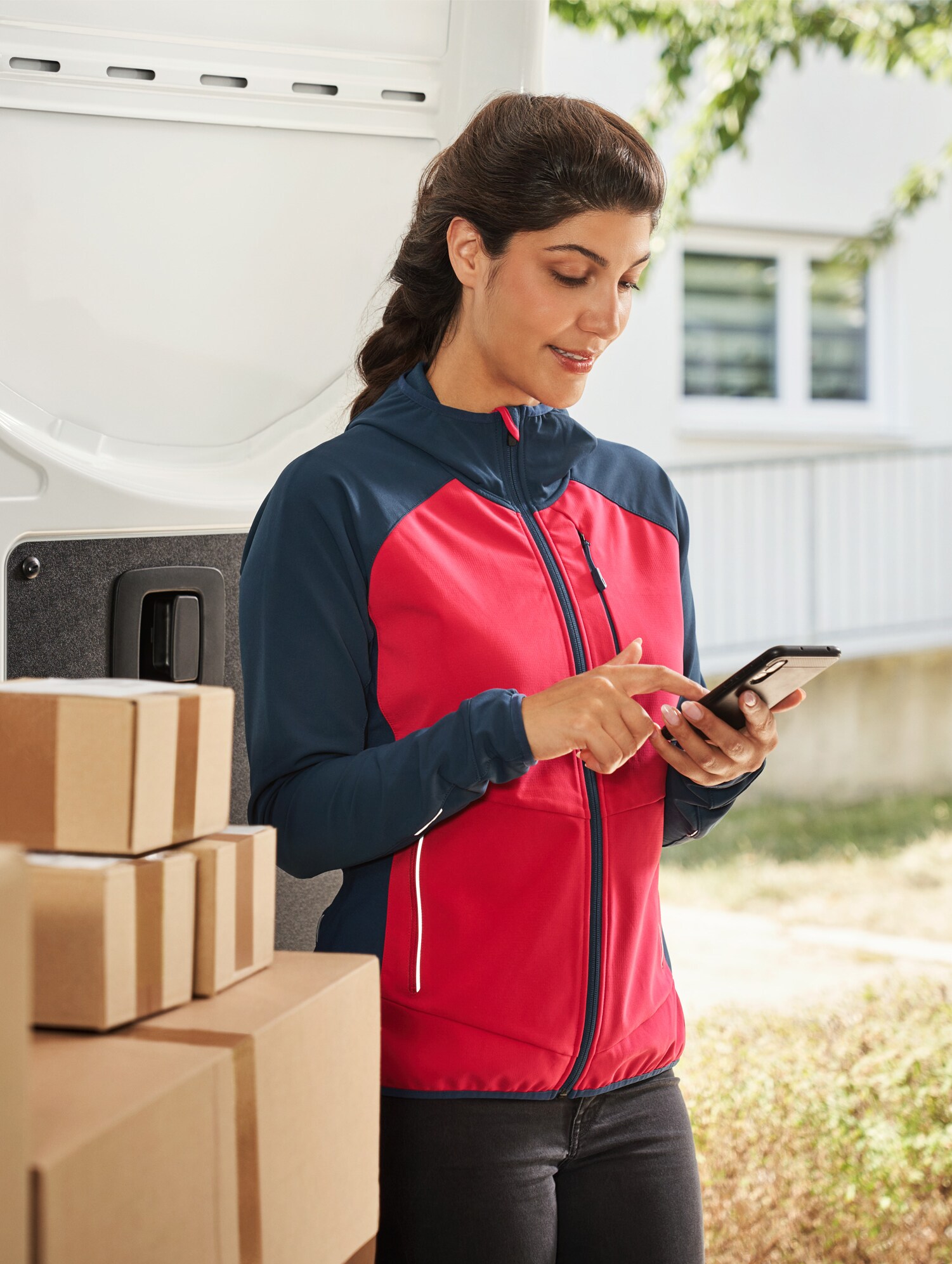 Delivery woman in front of a delivery van with parcels using a smartphone – professional delivery as part of Vans customer care.