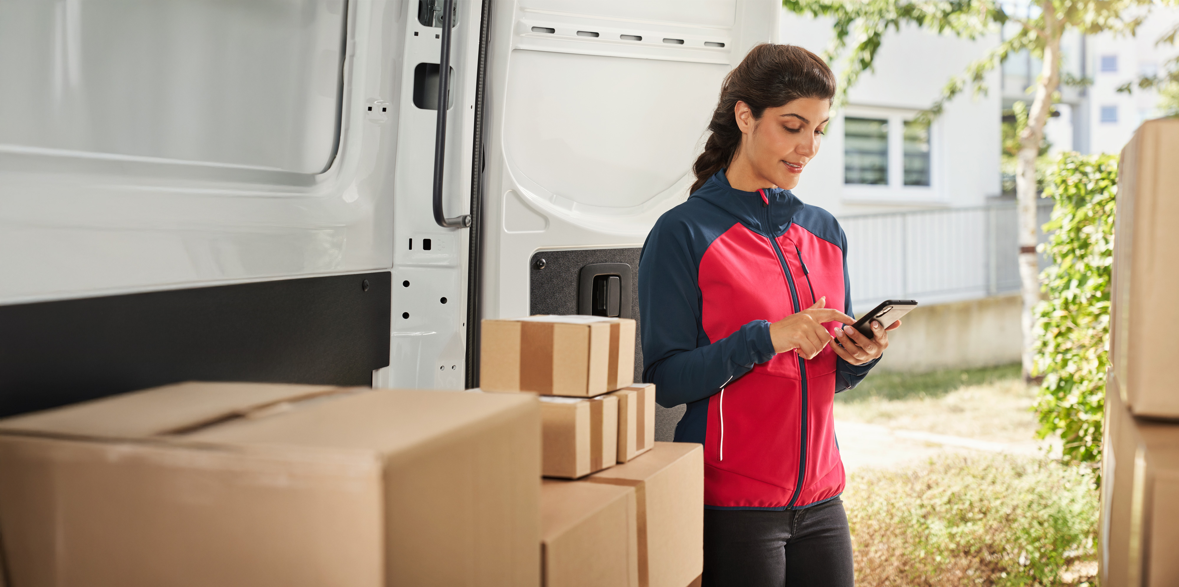Mercedes-Benz | Van care | Customer Assistance Center Delivery woman in front of a delivery van with parcels using a smartphone – professional delivery as part of Vans customer care.