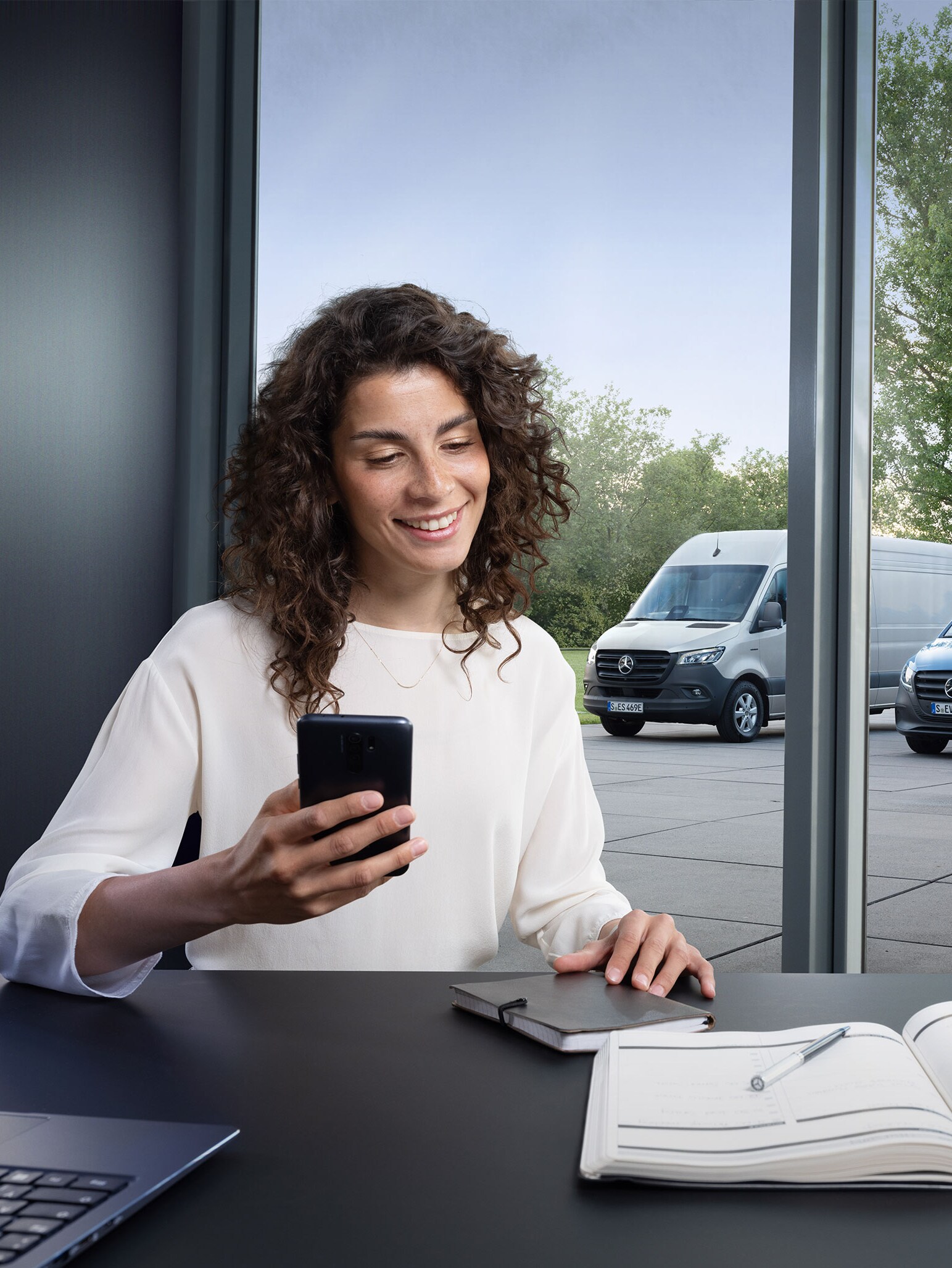 Woman with smartphone at a desk, serveral Mercedes-Benz vans in the background – expert support in business care.
