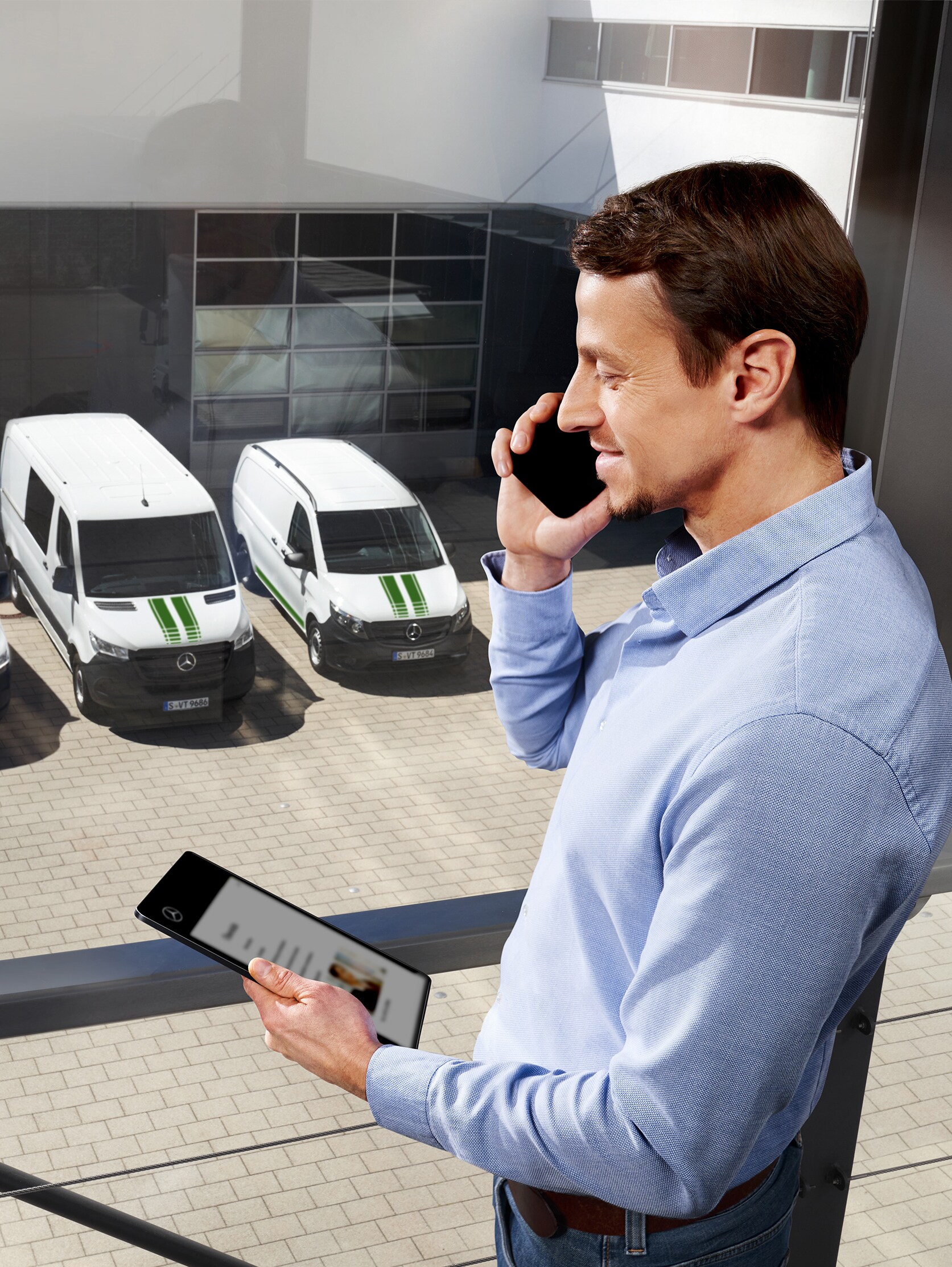 A man talking on a mobile phone in front of a small fleet of white Mercedes-Benz vans in a car park.