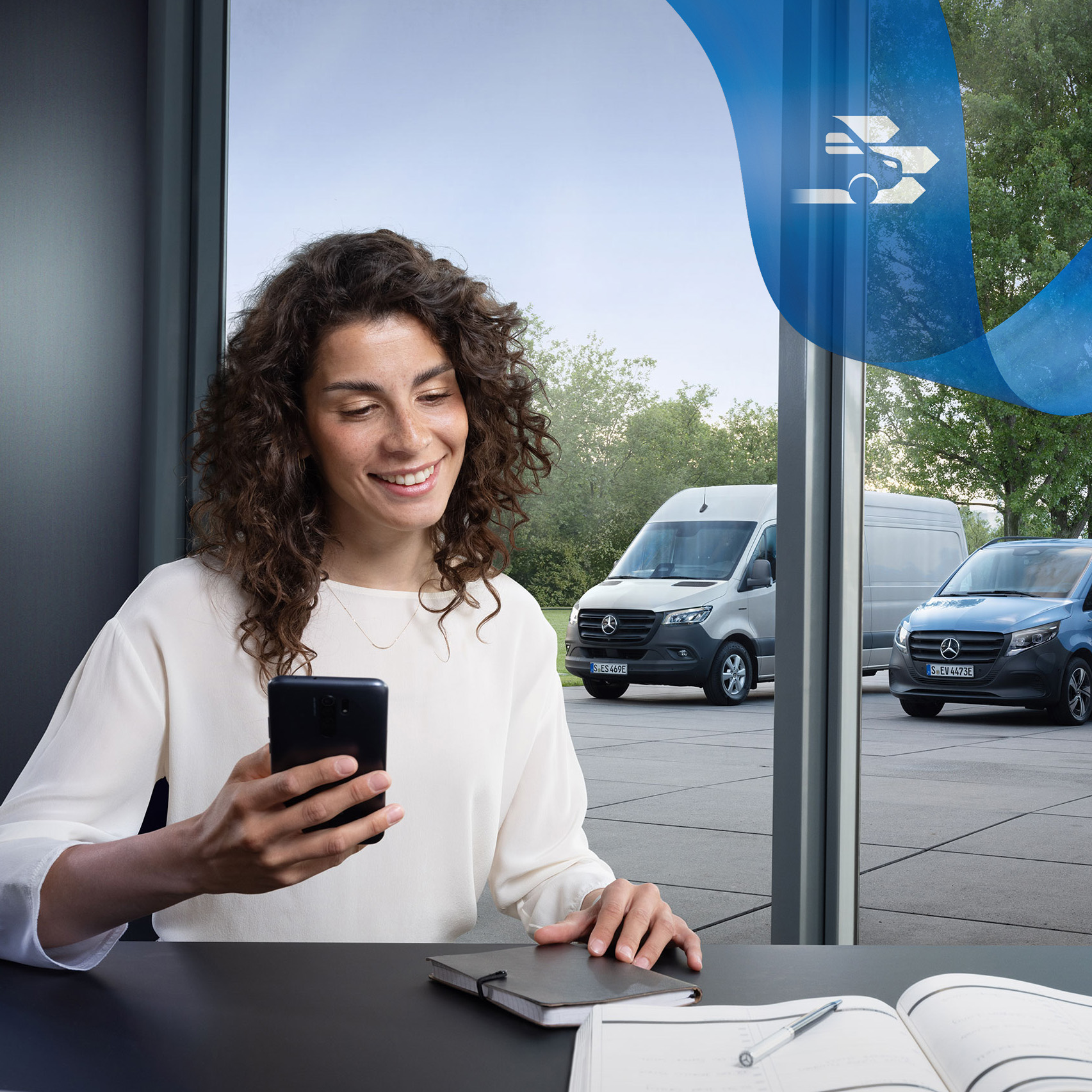 Mercedes-Benz | Van care | Customer Assistance Center Woman sitting at a desk with smartphone and a range of Mercedes-Benz vans in the background.