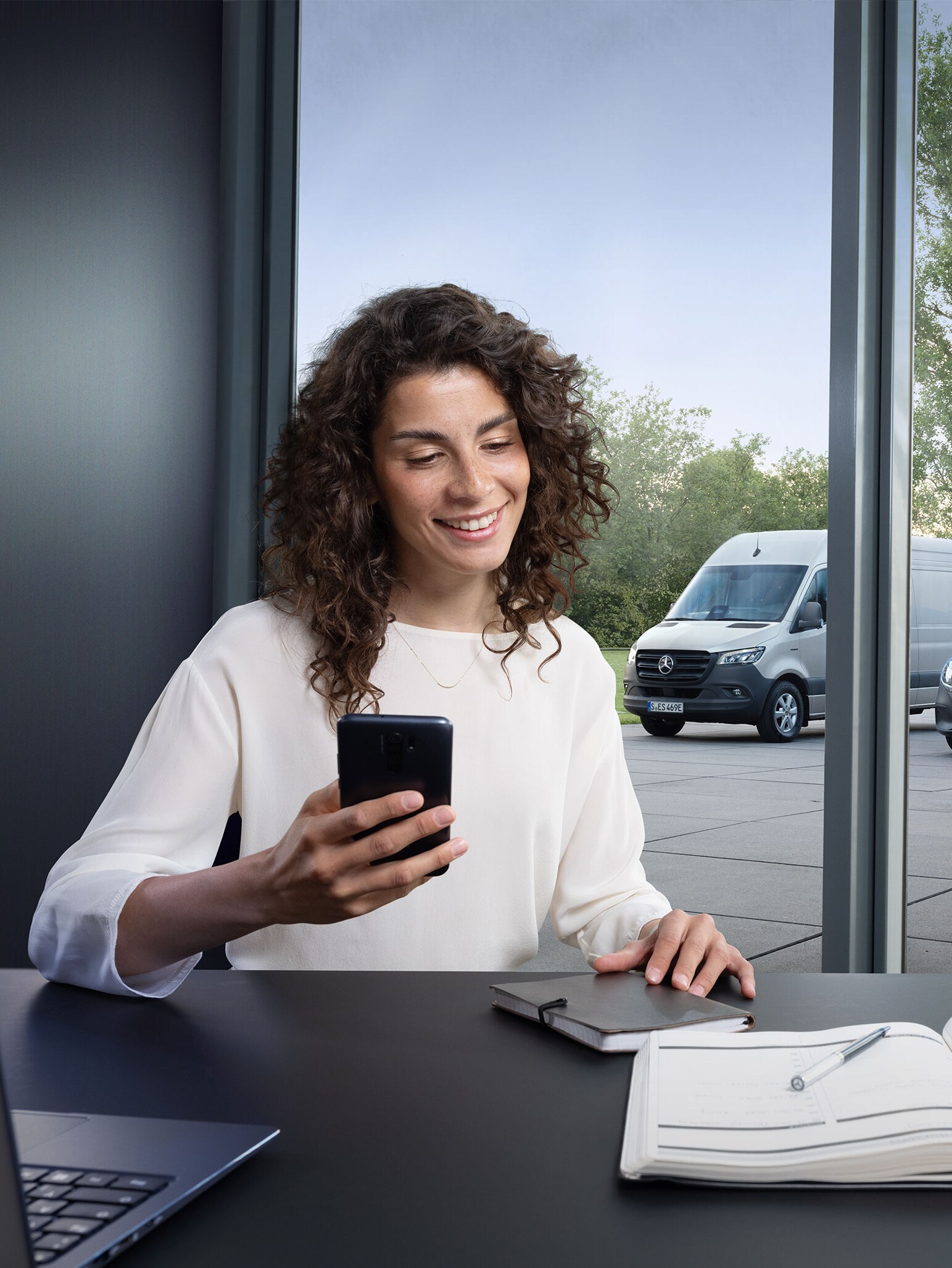 Woman booking a service via smartphone with a range of Mercedes-Benz vans in the background.