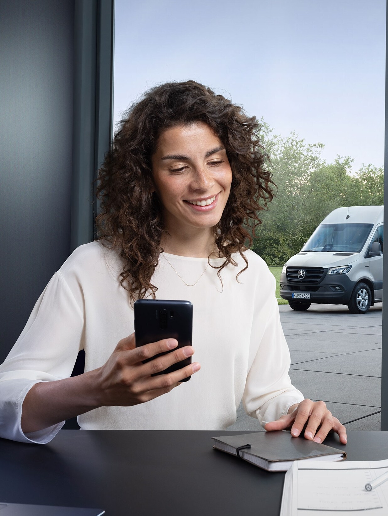 Woman using a smartphone to control the fleet monitoring of several Mercedes-Benz vans at the office window.
