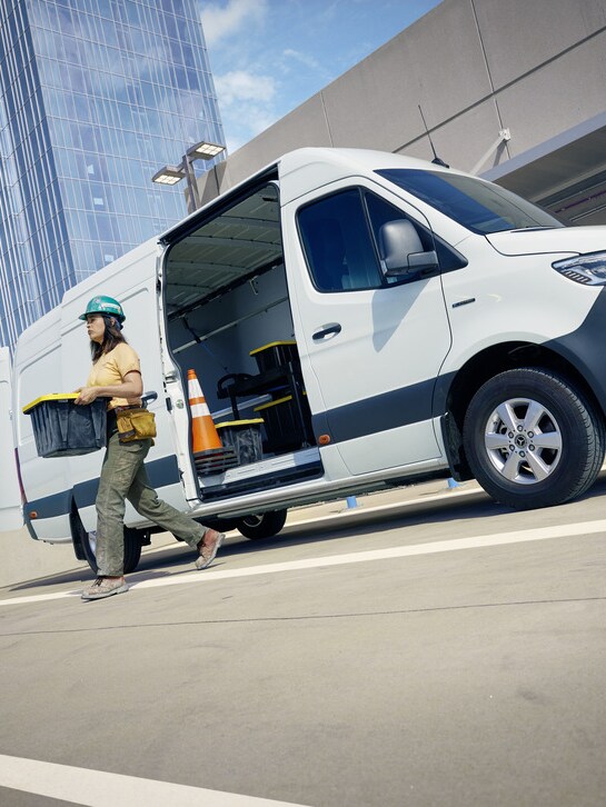 A person carrying a box next to a white Mercedes-Benz eSprinter Panel van.