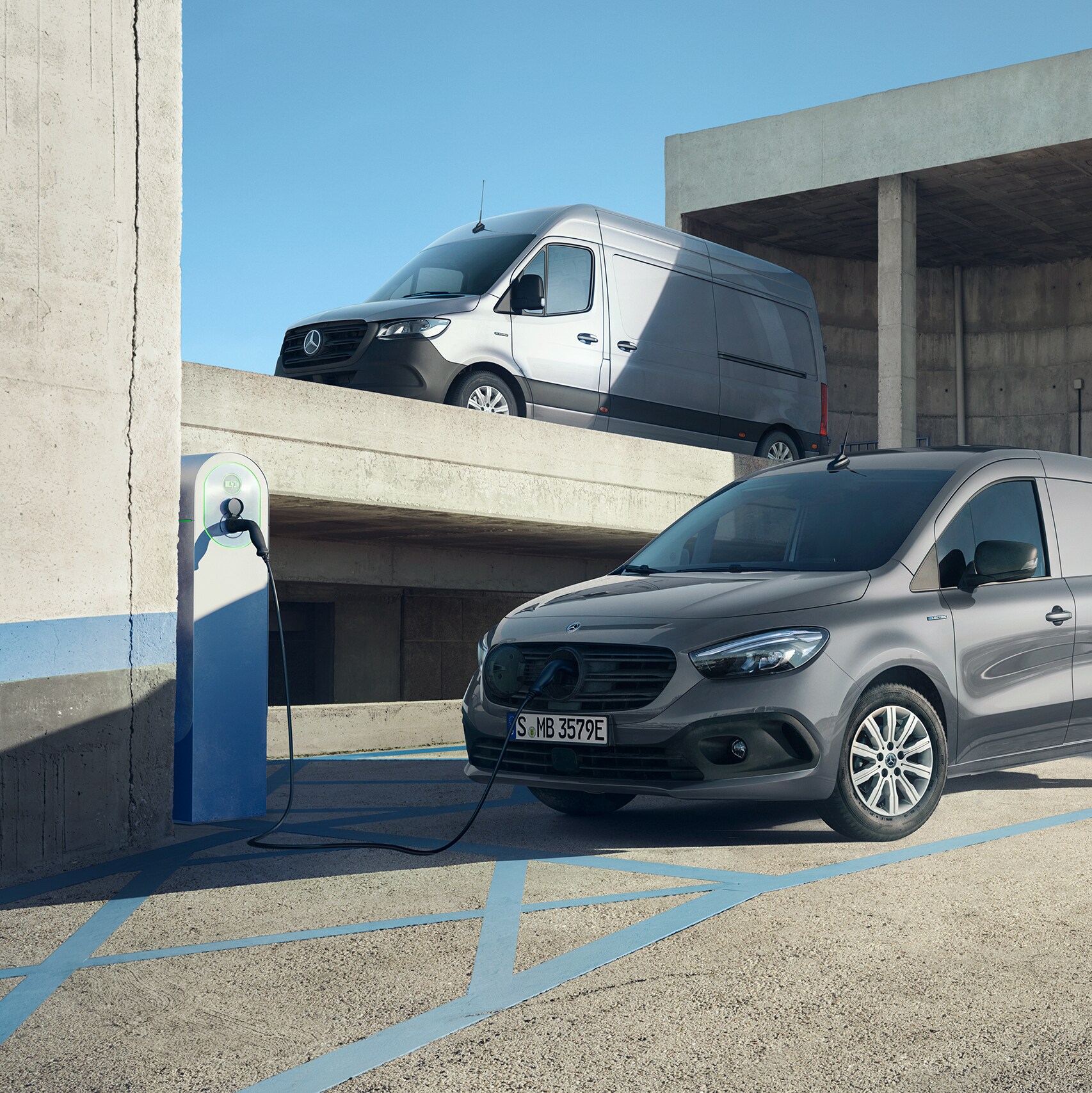 A light grey Mercedes-Benz eSprinter Panel van and a dark grey Mercedes-Benz eCitan van in an industrial setting, with the eCitan van charging at a charging station.