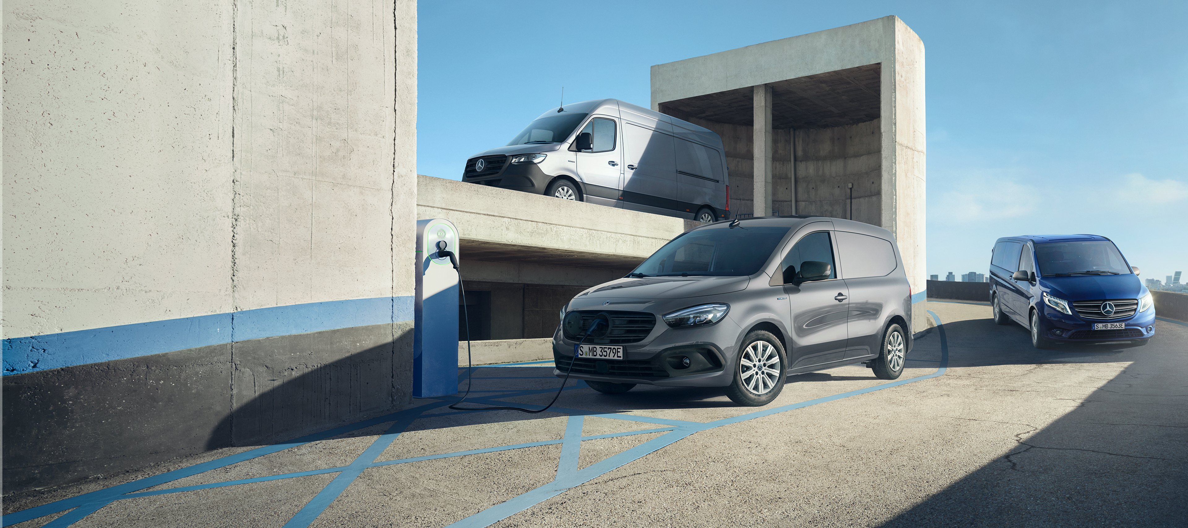 Grey eSprinter Panel Van & eCitan Van | Mercedes-Benz Vans A light grey Mercedes-Benz eSprinter Panel van and a dark grey Mercedes-Benz eCitan van in an industrial setting, with the eCitan van charging at a charging station.