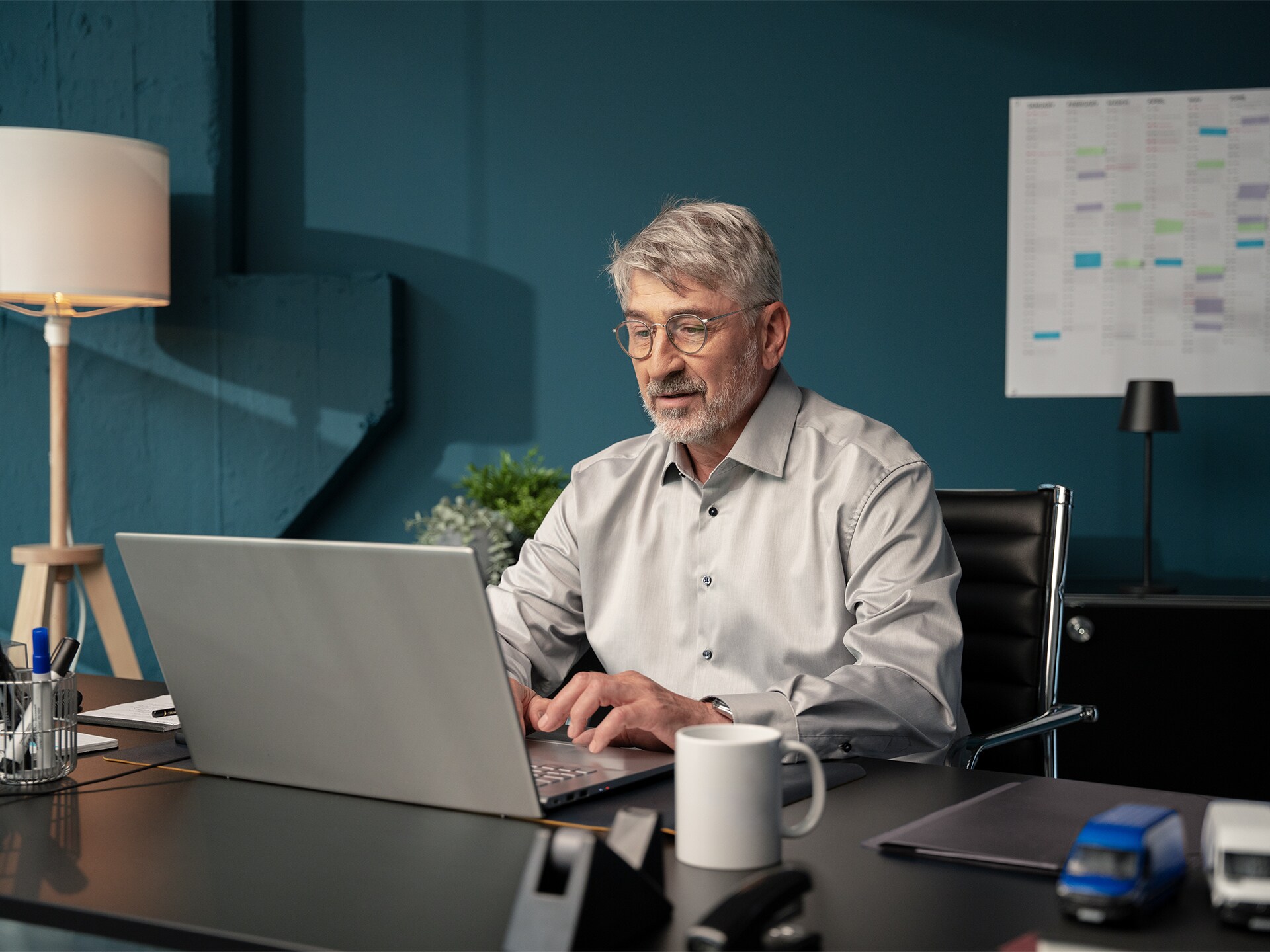 A man with glasses and a grey shirt working on a silver laptop at a desk in an office.