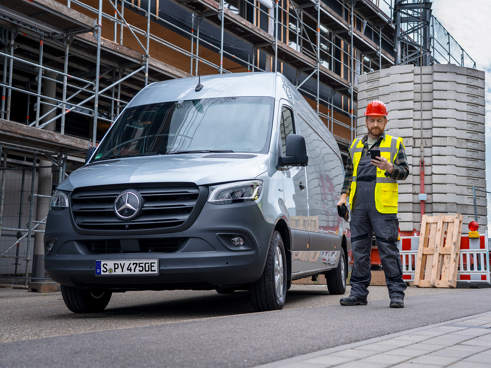 Silver Sprinter van at construction site | Mercedes-Benz Vans A construction worker in a red hard hat and high vis vest looking at a phone next to a silver Mercedes-Benz van parked at a building site.