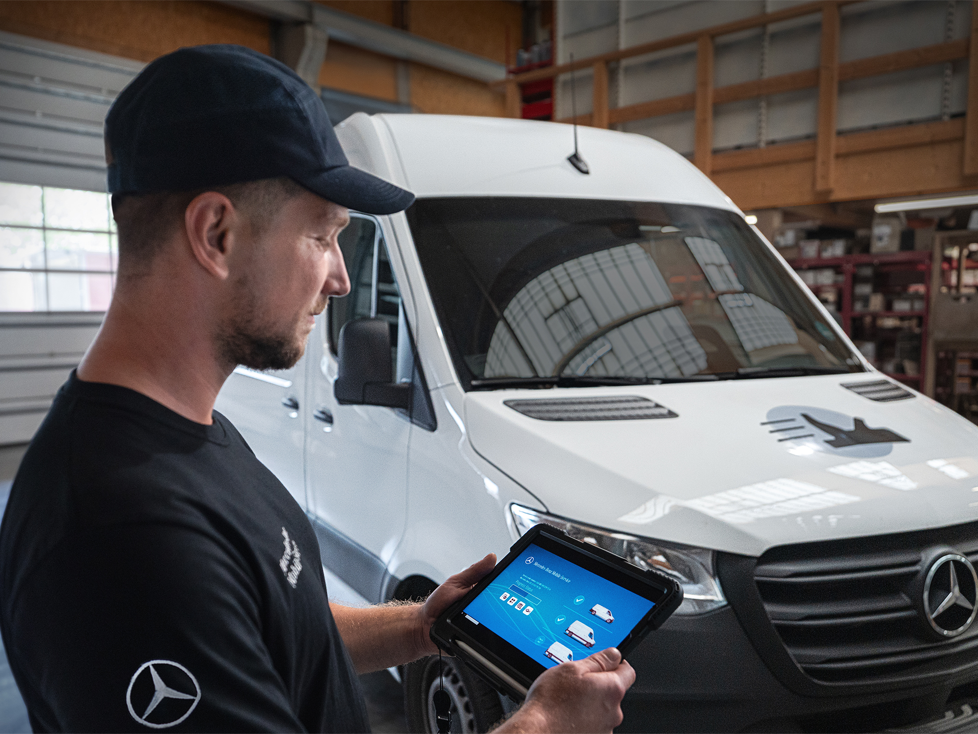 Technician with tablet | Mercedes-Benz Vans A technician in a black cap and shirt looking at a tablet, with a white Mercedes-Benz van parked behind him in a workshop.