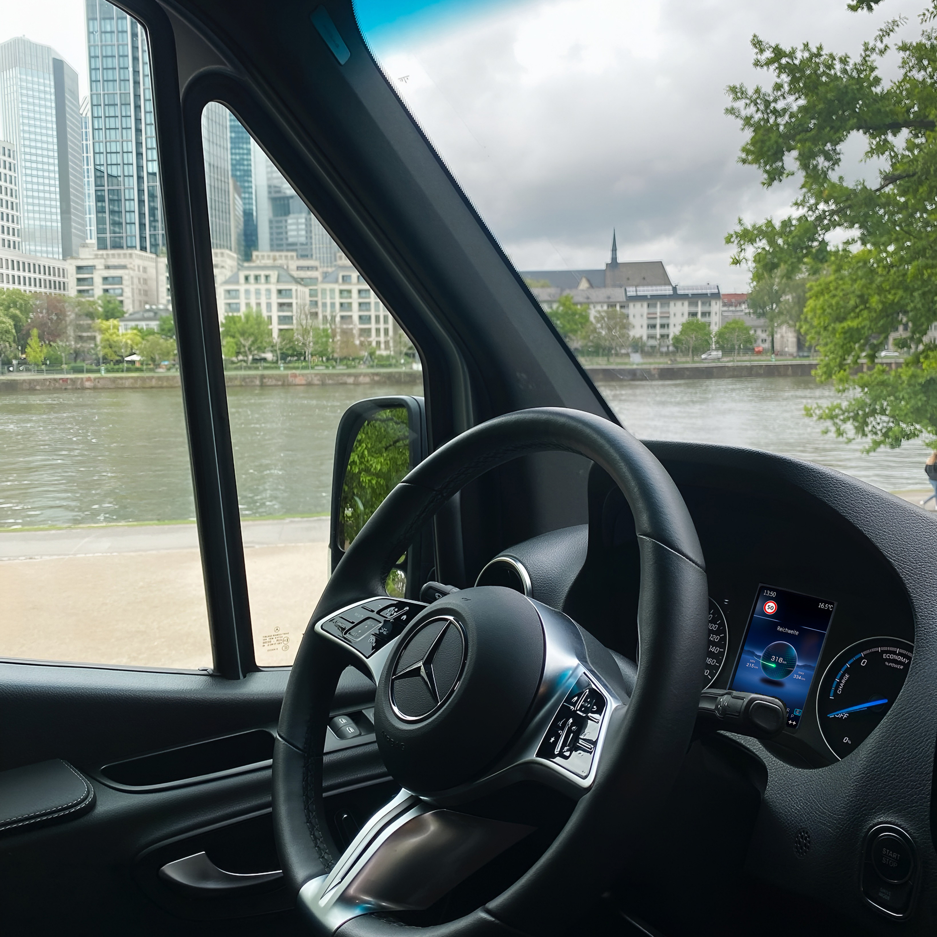 The Interior of a Mercedes-Benz Van Overlooking a City River | Mercedes-Benz Vans A view from the driver's seat showing the steering wheel and a city river and skyline through the window.