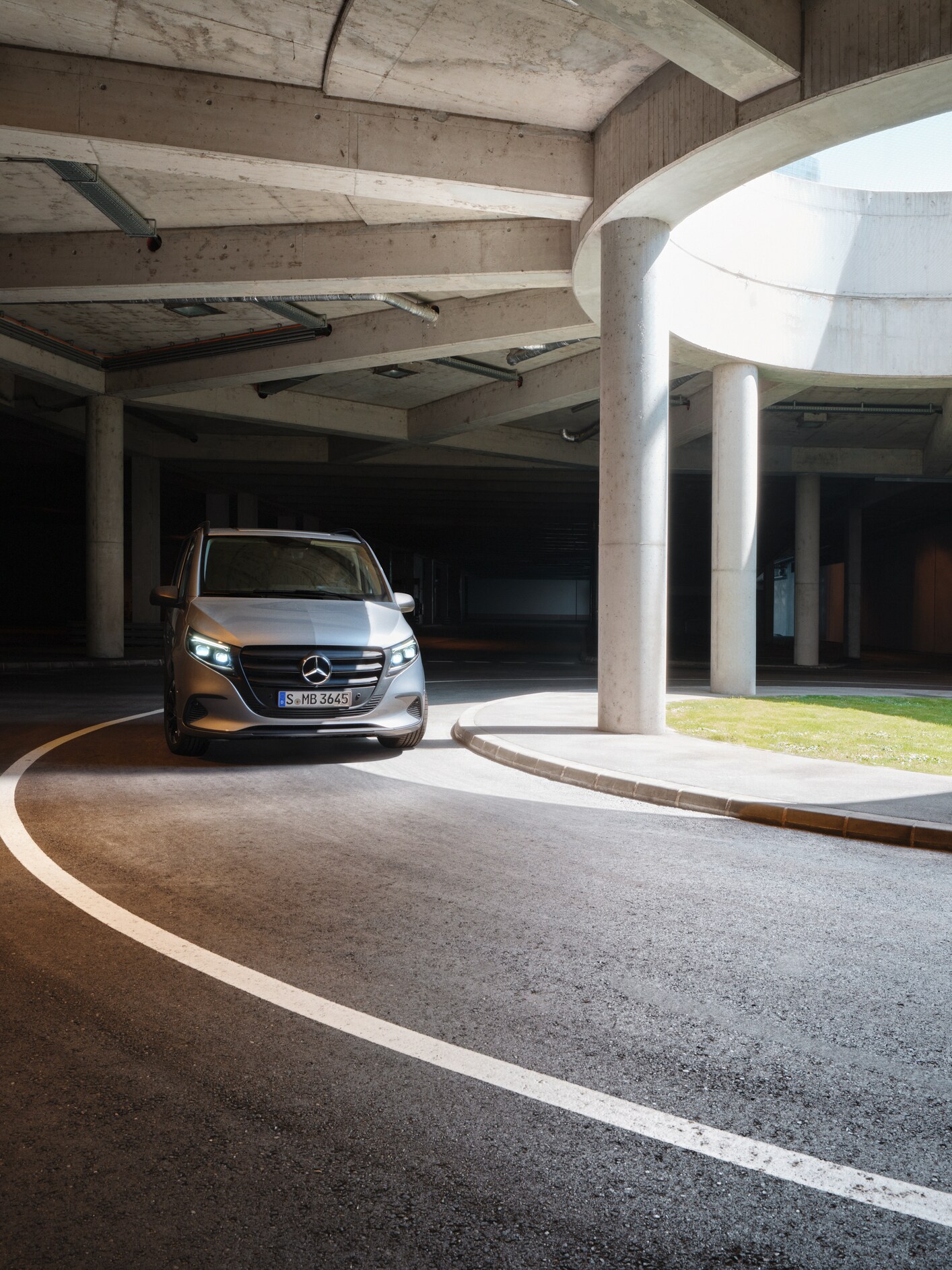 A silver Mercedes-Benz van driving through a semi-covered car park with concrete pillars and a grass verge.