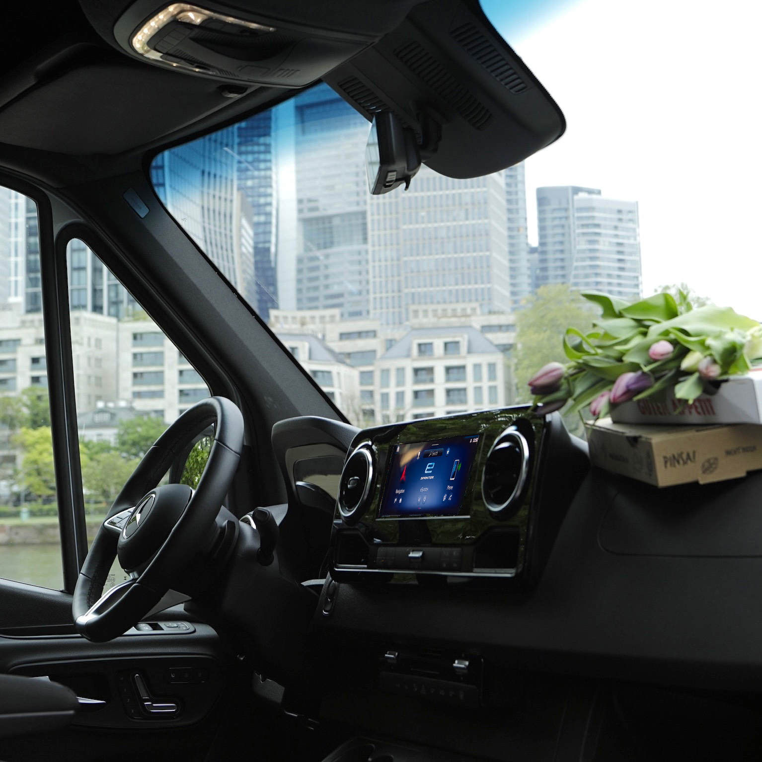 The Interior of a Mercedes-Benz Van in a City | Mercedes-Benz Vans The cabin interior of a Mercedes-Benz van showing the dashboard, steering wheel, and a city skyline through the windscreen.