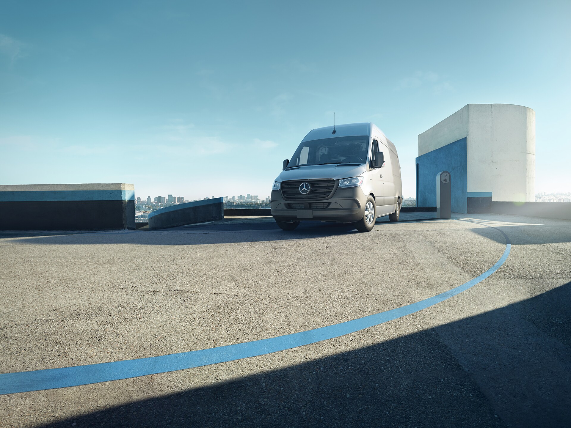 Grey Mercedes-Benz van parked distant city skyline under a clear blue sky on an open rooftop parking deck beside a modern charging station.