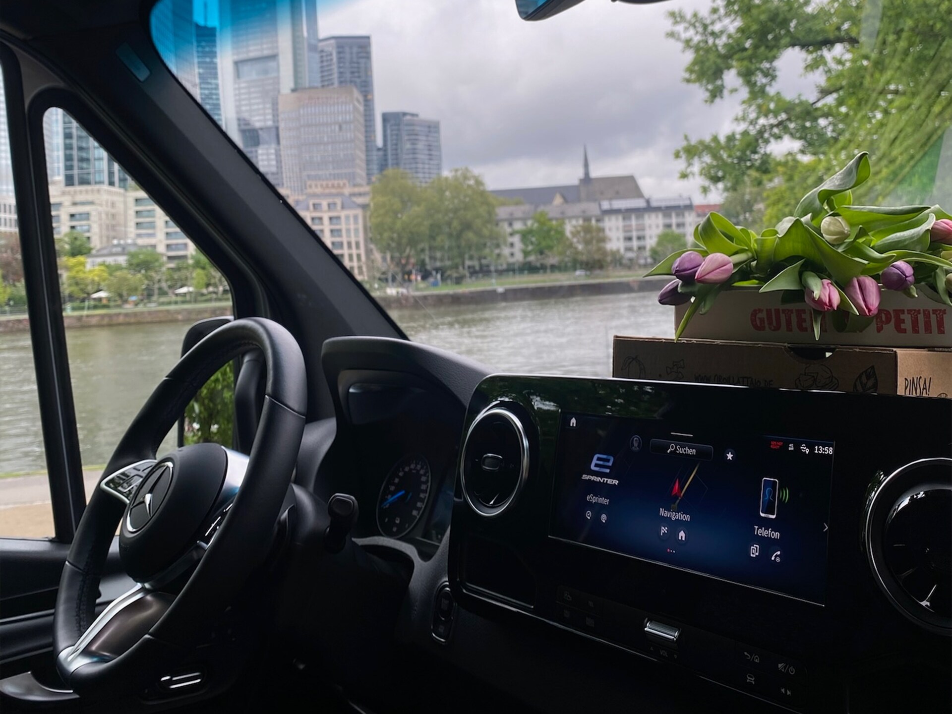 The interior of a Mercedes-Benz vehicle focusing on dashboard and steering wheel with cityscape and river visible through window.