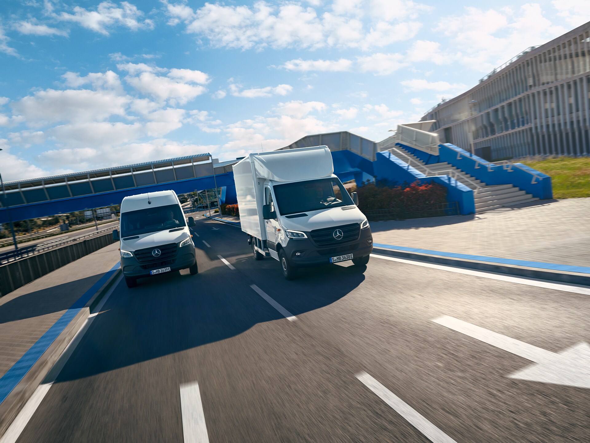 Two white Mercedes-Benz vans driving side by side on a multi-lane road under a blue pedestrian bridge with stairs.