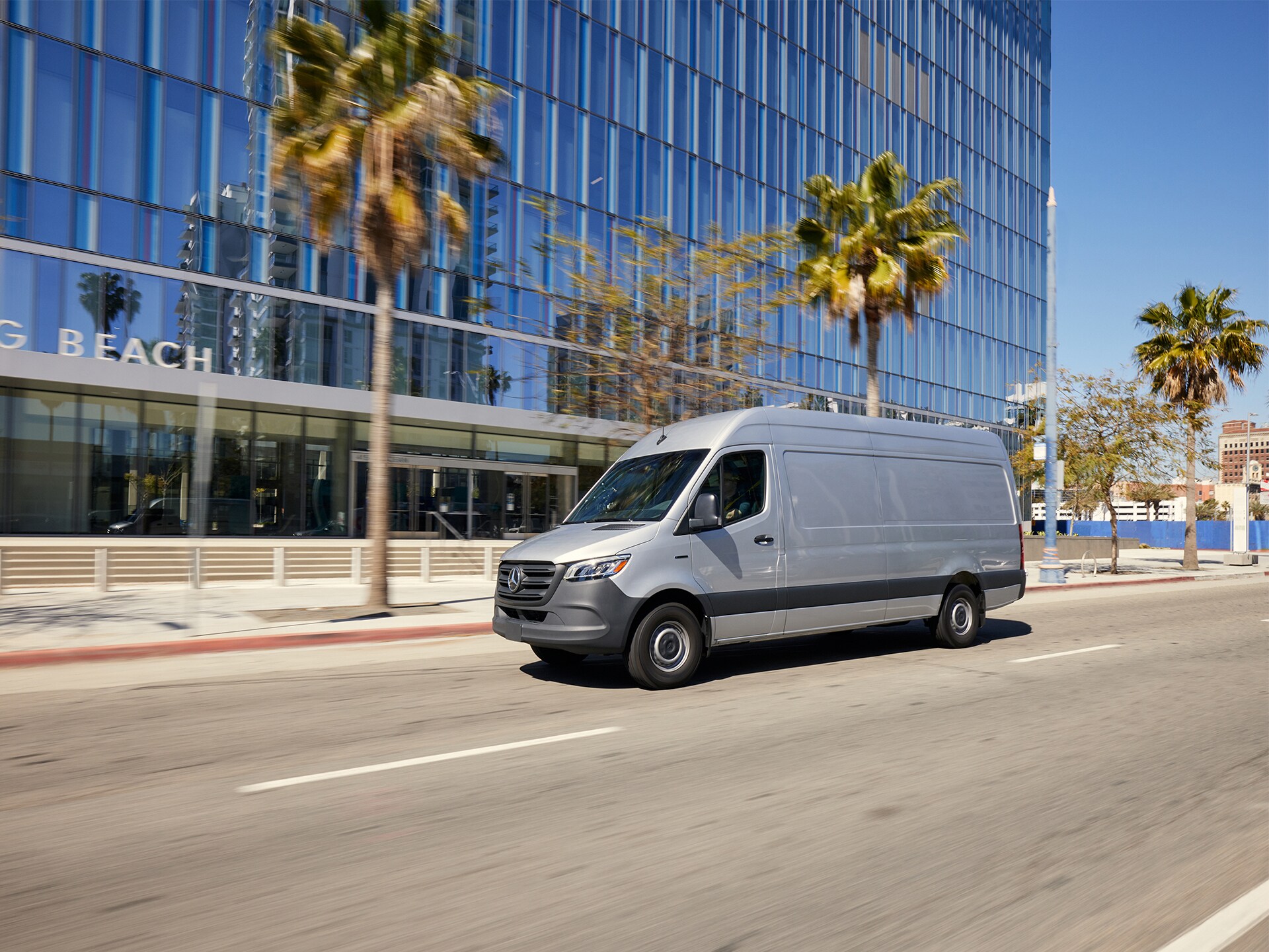 Silver Sprinter van driving on a city street lined with palm trees and modern glass buildings.
