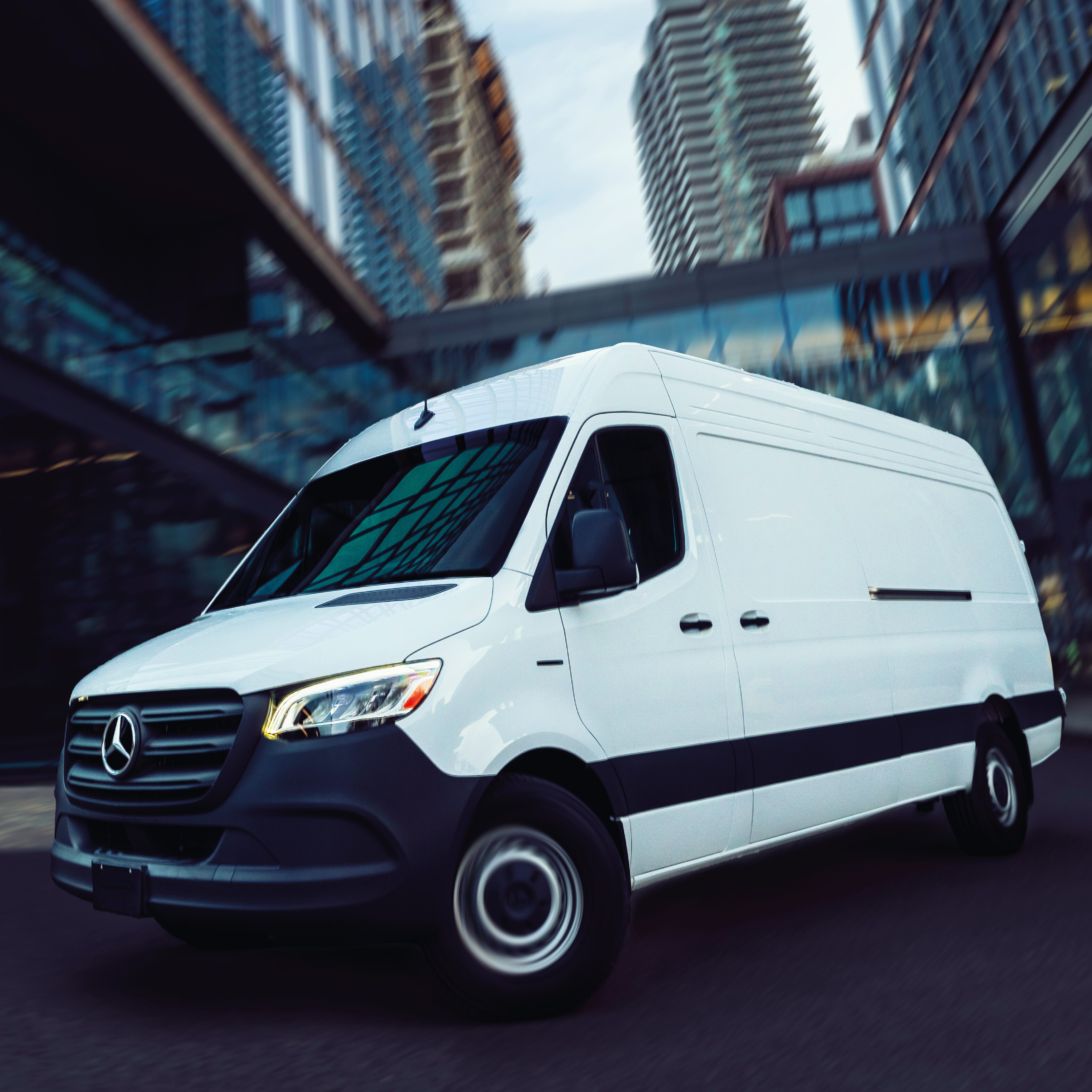 White Mercedes-Benz Sprinter delivery van driving through an urban area with modern glass buildings and high-rises in background