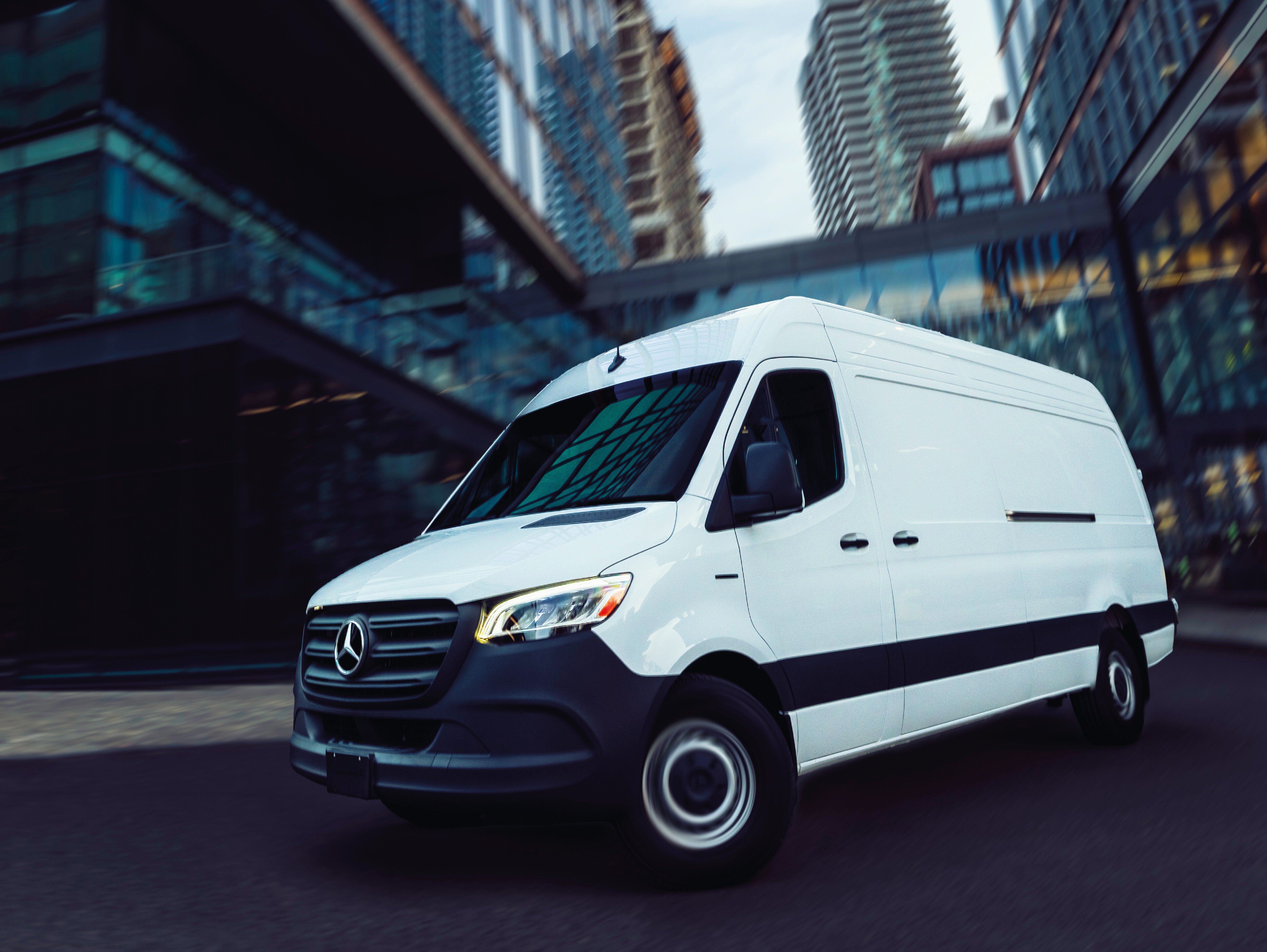 White Mercedes-Benz Sprinter delivery van driving through an urban area with modern glass buildings and high-rises in background