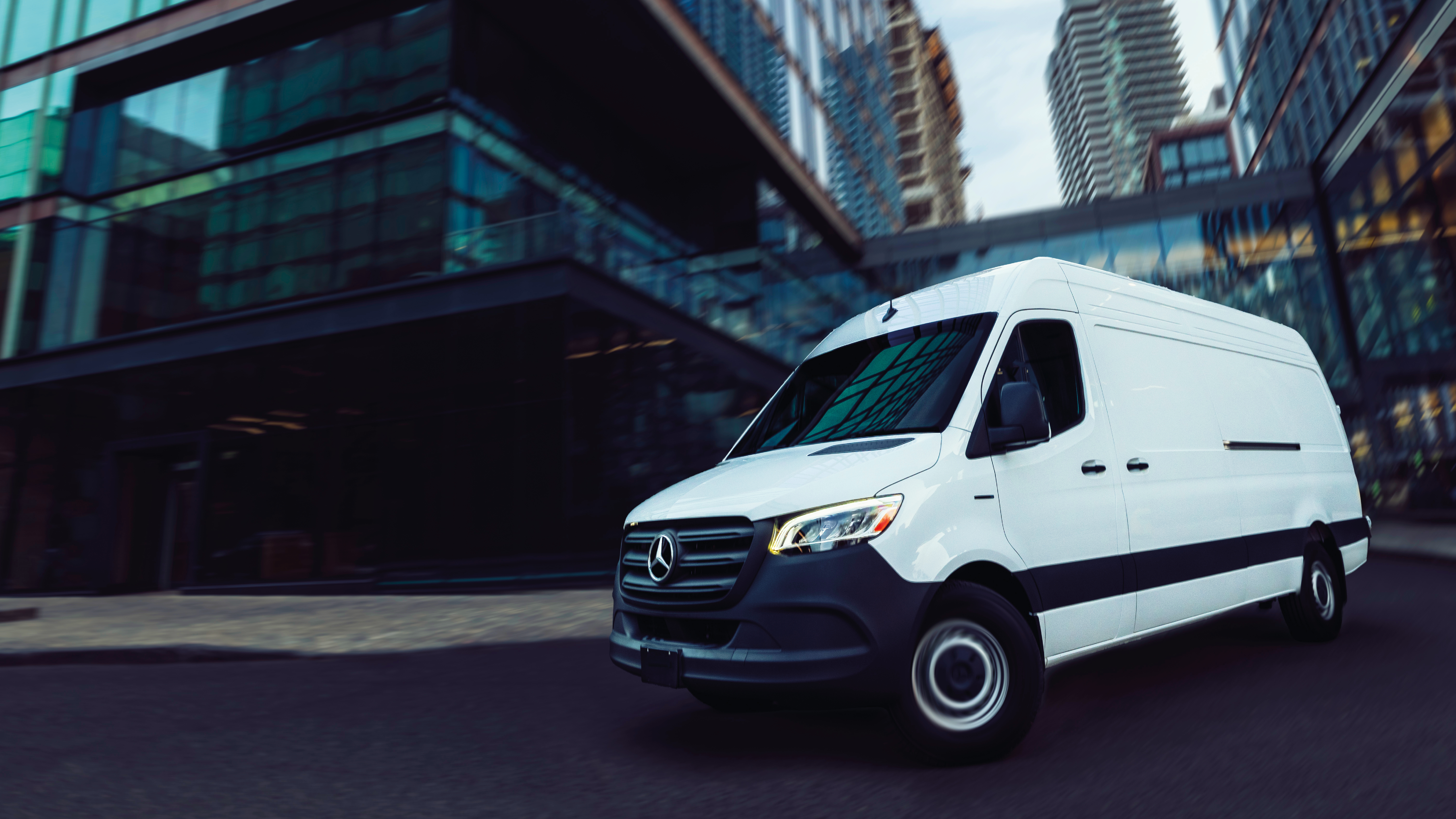 Sprinter delivery van in urban setting | Mercedes-Benz Vans White Mercedes-Benz Sprinter delivery van driving through an urban area with modern glass buildings and high-rises in background