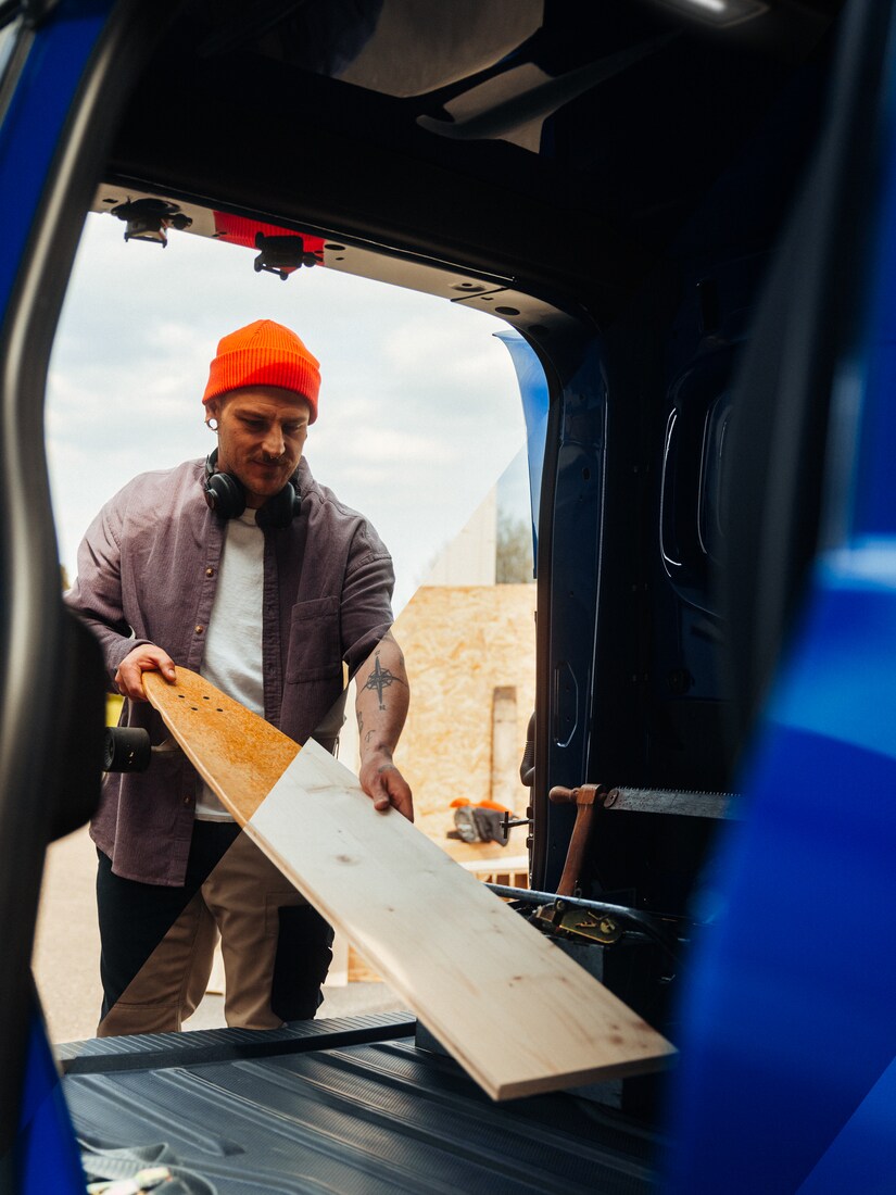 A man in an orange beanie and headphones loading a long, flat wooden board into the back of a blue Mercedes-Benz van.