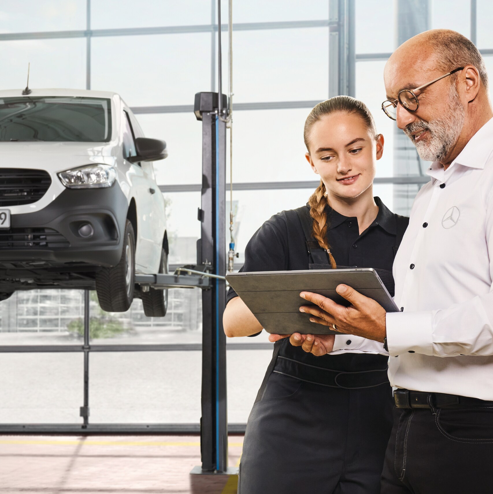 Two Mercedes-Benz mechanics stand in a garage discussing in front of a tablet beside a hoisted van.