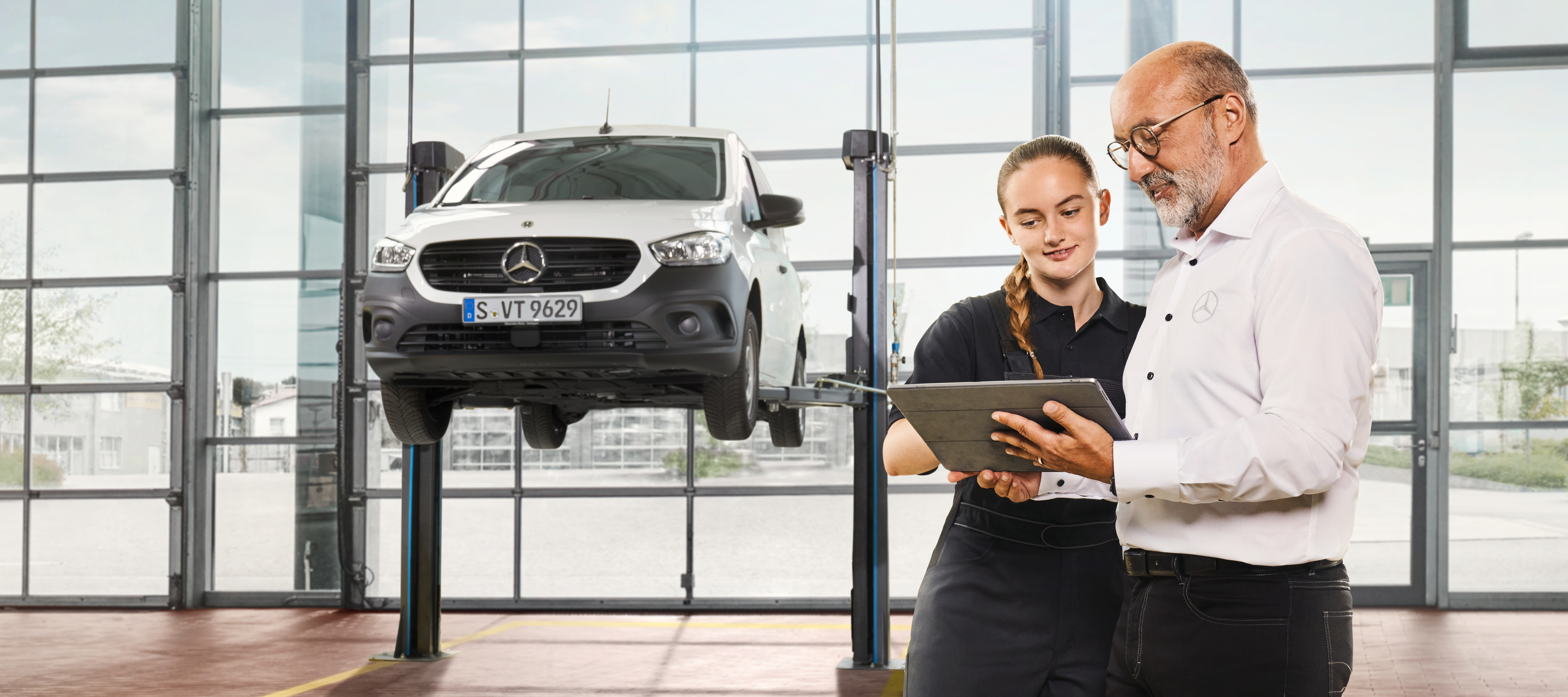 Technicians Reviewing a Tablet | Mercedes-Benz Vans Two Mercedes-Benz mechanics stand in a garage discussing in front of a tablet beside a hoisted van.