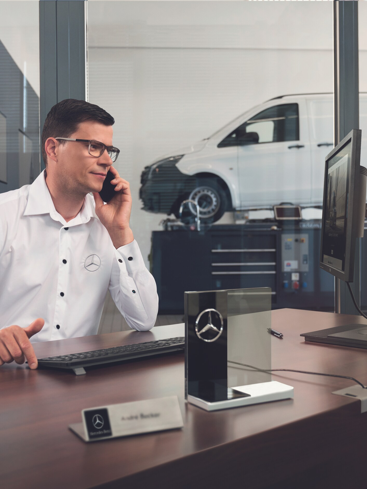 A Mercedes-Benz service advisor in glasses talking on the phone at his desk with a white van in the background.