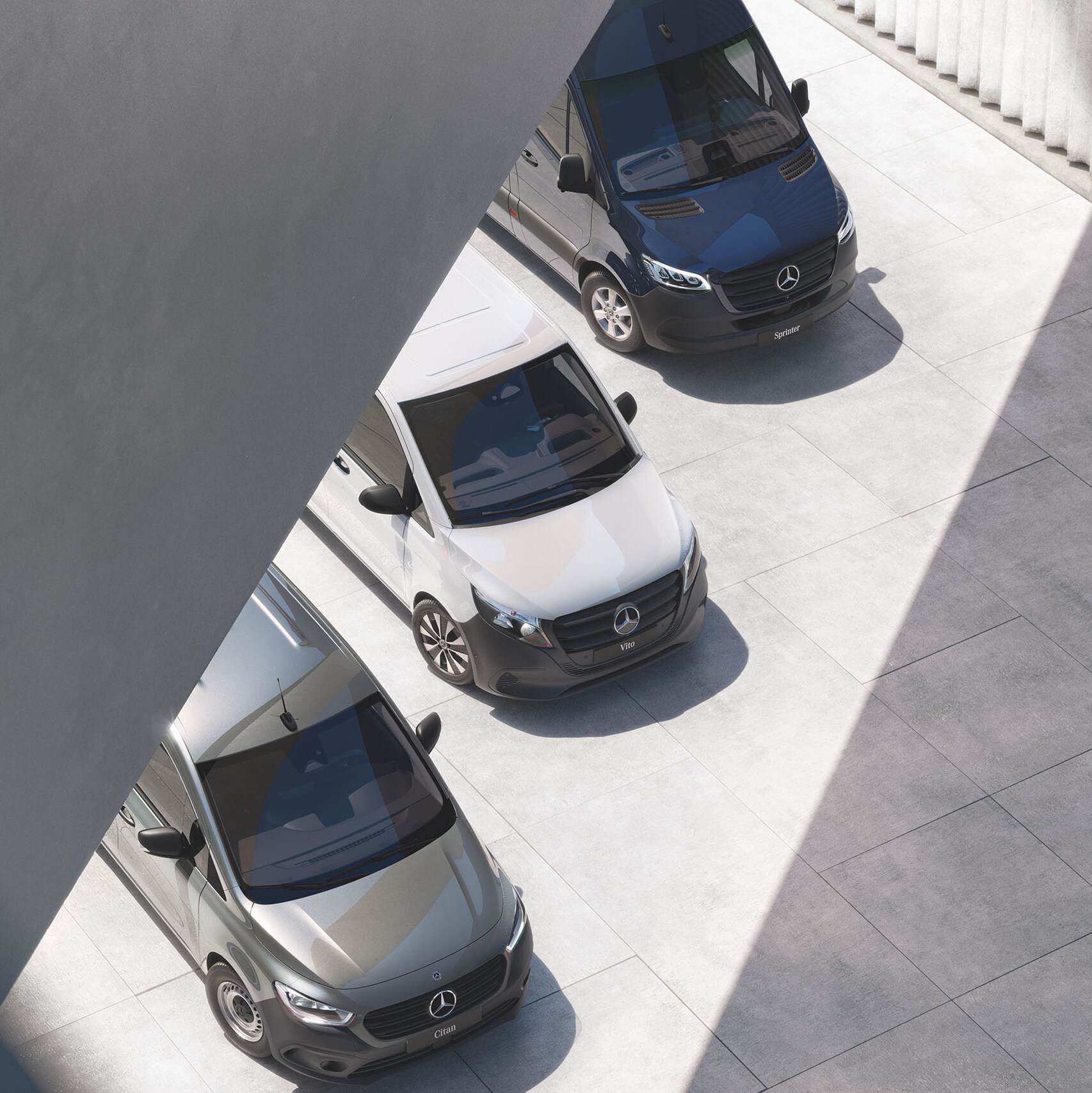Overhead view of Mercedes-Benz vans in blue, white, grey, parked diagonally in sunlit industrial courtyard by metal wall.
