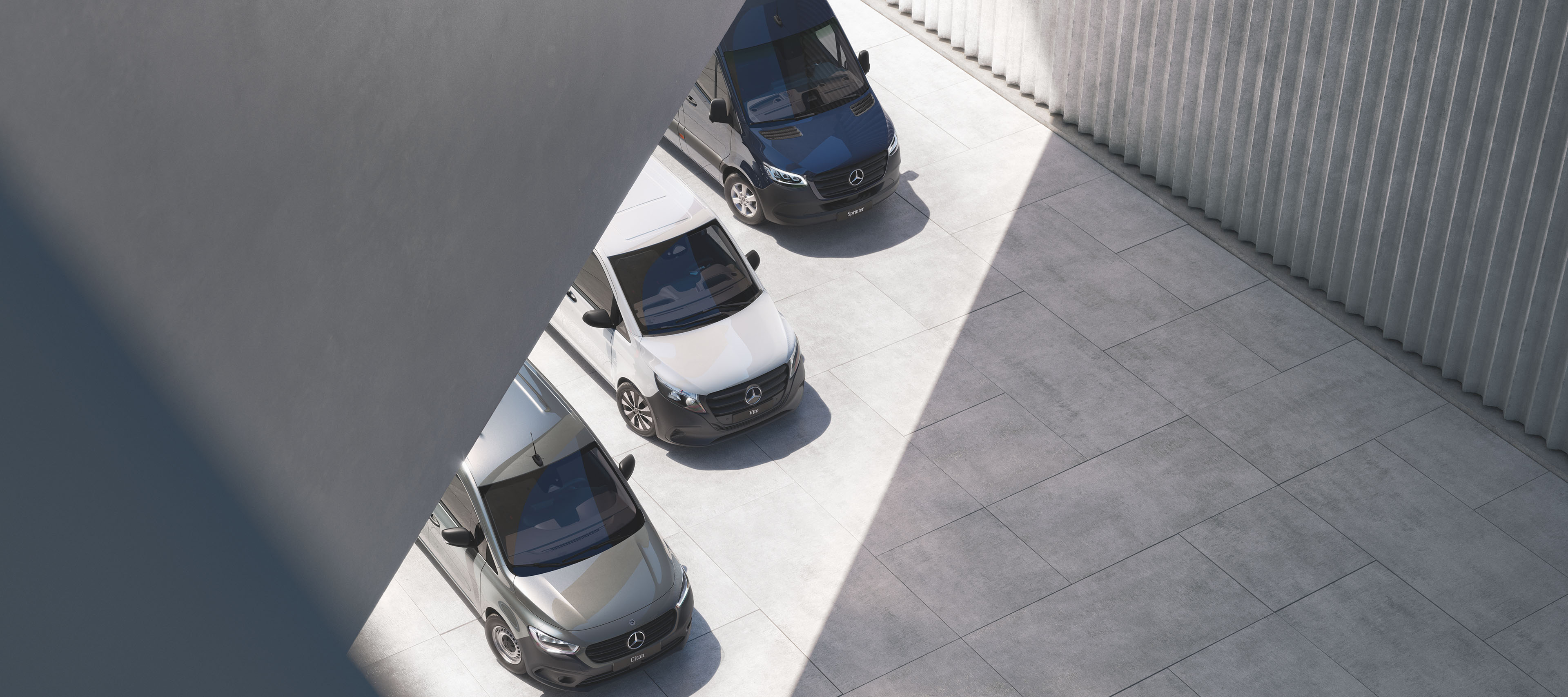 Mercedes-Benz Vans commercial range Overhead view of Mercedes-Benz vans in blue, white, grey, parked diagonally in sunlit industrial courtyard by metal wall.