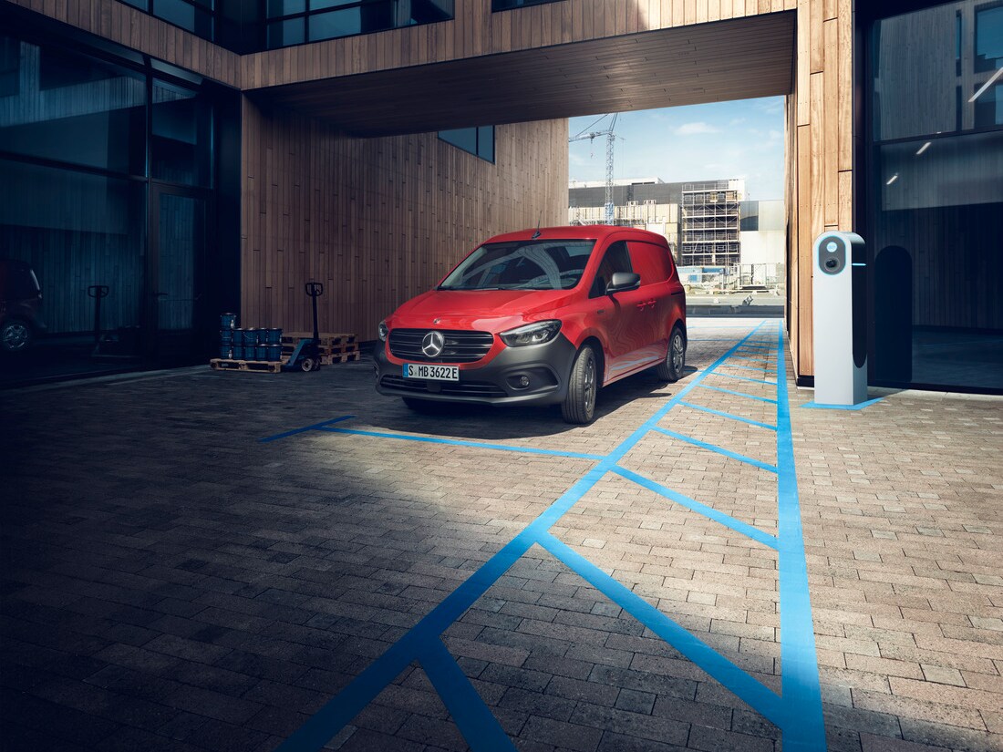 Red Mercedes-Benz eCitan Panel Van parked in a charging bay outside a modern building.