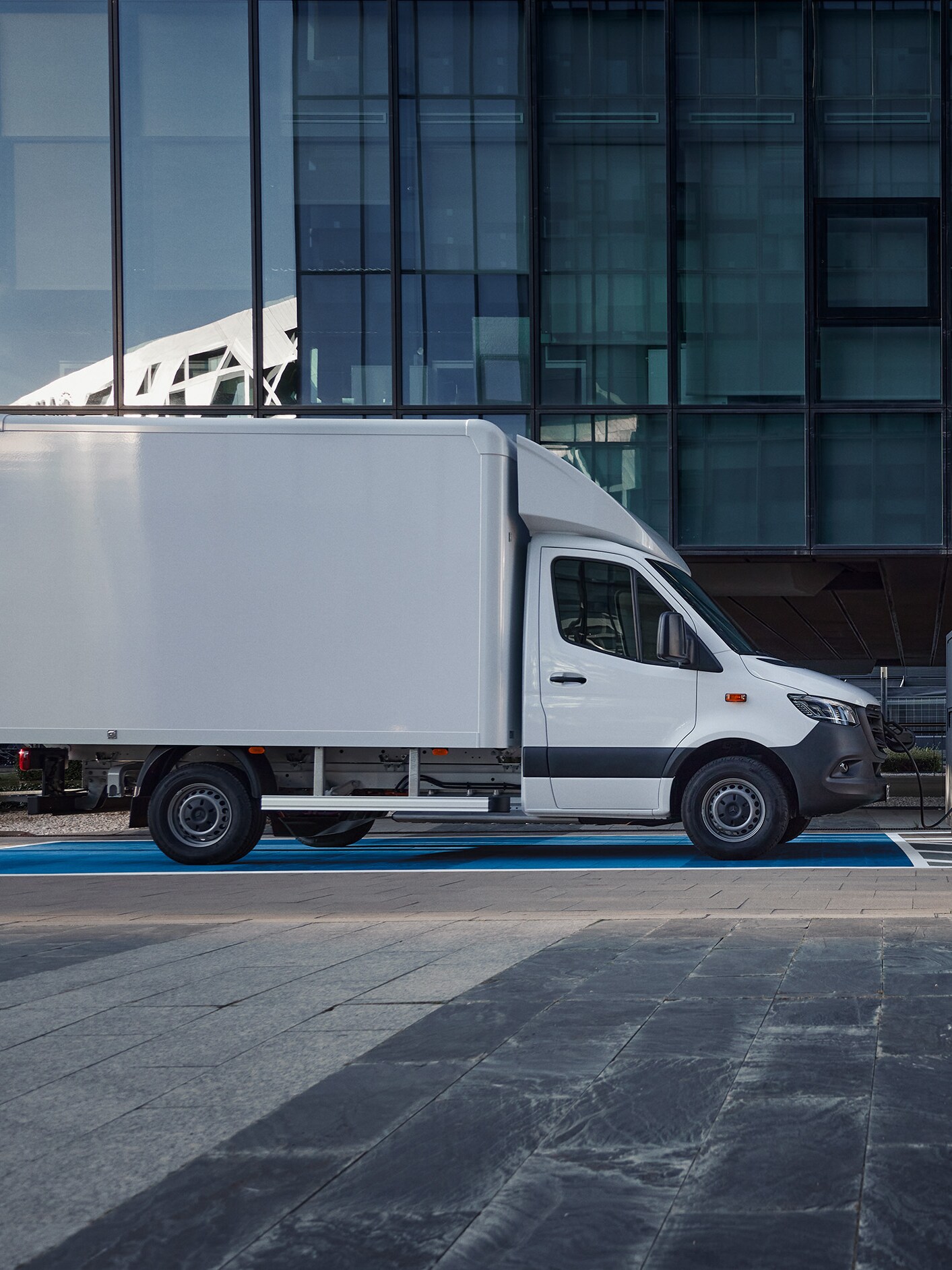 A white Mercedes-Benz eSprinter Luton Box van plugged into an electric charger in front of a building.