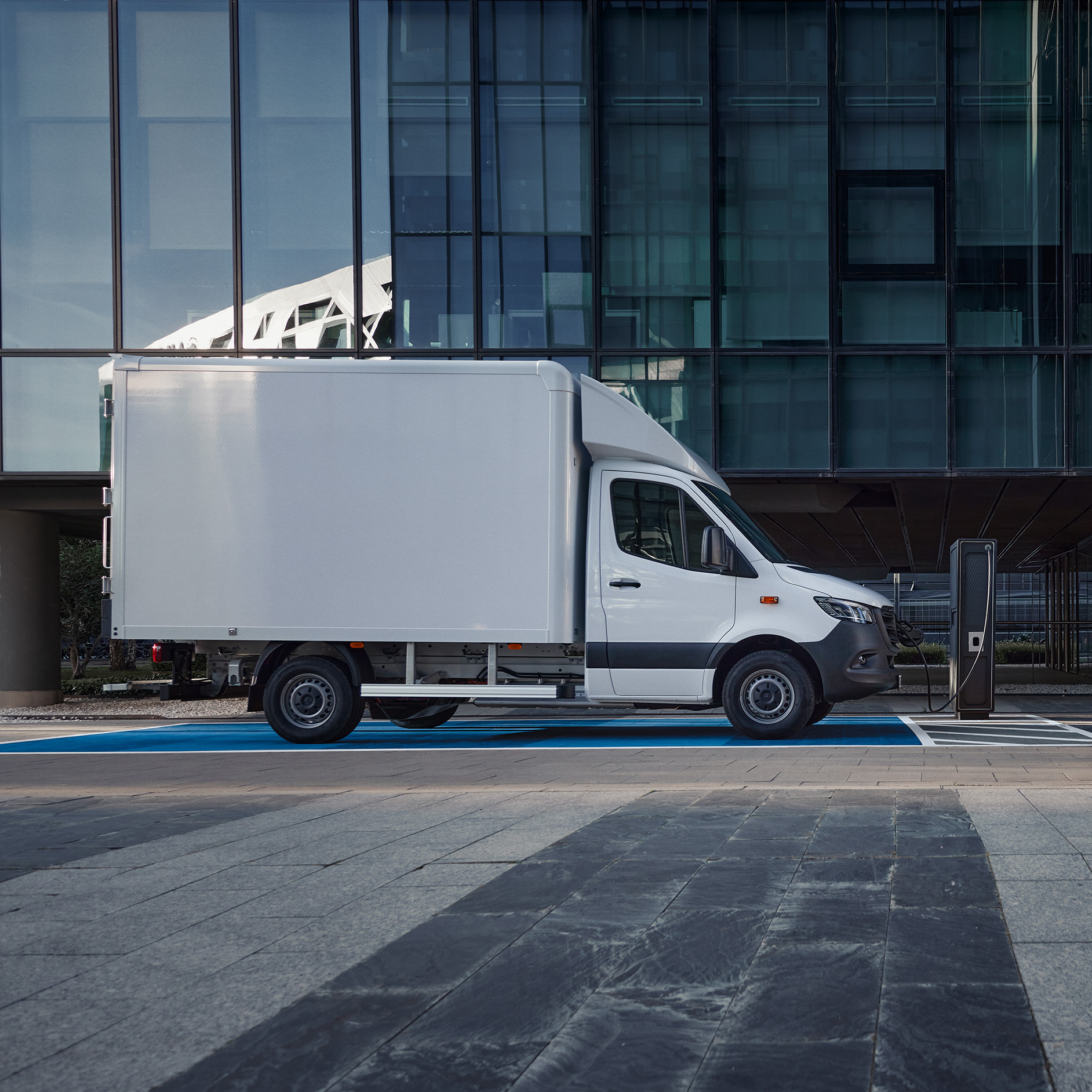 eSprinter Luton Box Van Charging | Mercedes-Benz Vans A white Mercedes-Benz eSprinter Luton Box van plugged into an electric charger in front of a building.