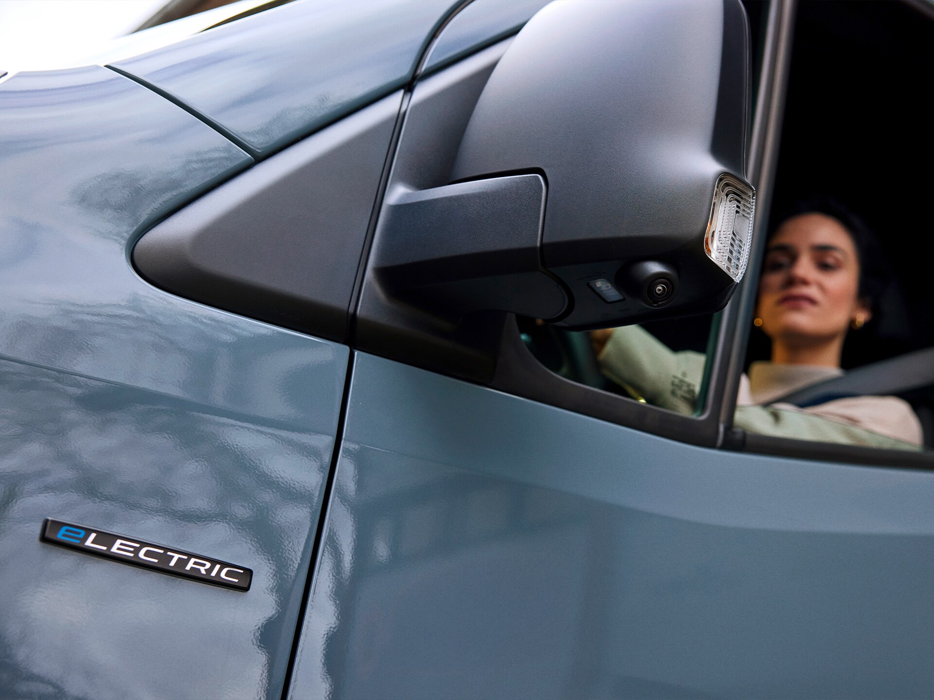 Close-up of a blue electric vehicle showing the side mirror, "electric" badge on the front door, and part of the driver visible through the window.